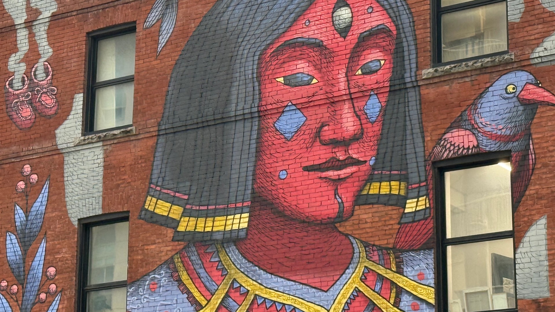 SbuOne mural in Lowertown Ottawa featuring a red-faced, blue-eyed figure with a third eye and dark hair, wearing stylized armor, and a blue and red bird perched on its shoulder, painted on a red brick wall.