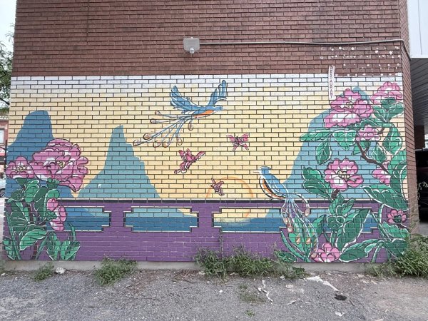 A mural on a brick wall depicts a blue bird, pink butterflies, and large flowers against blue mountains at 299 Bronson Avenue, Chinatown, Ottawa.