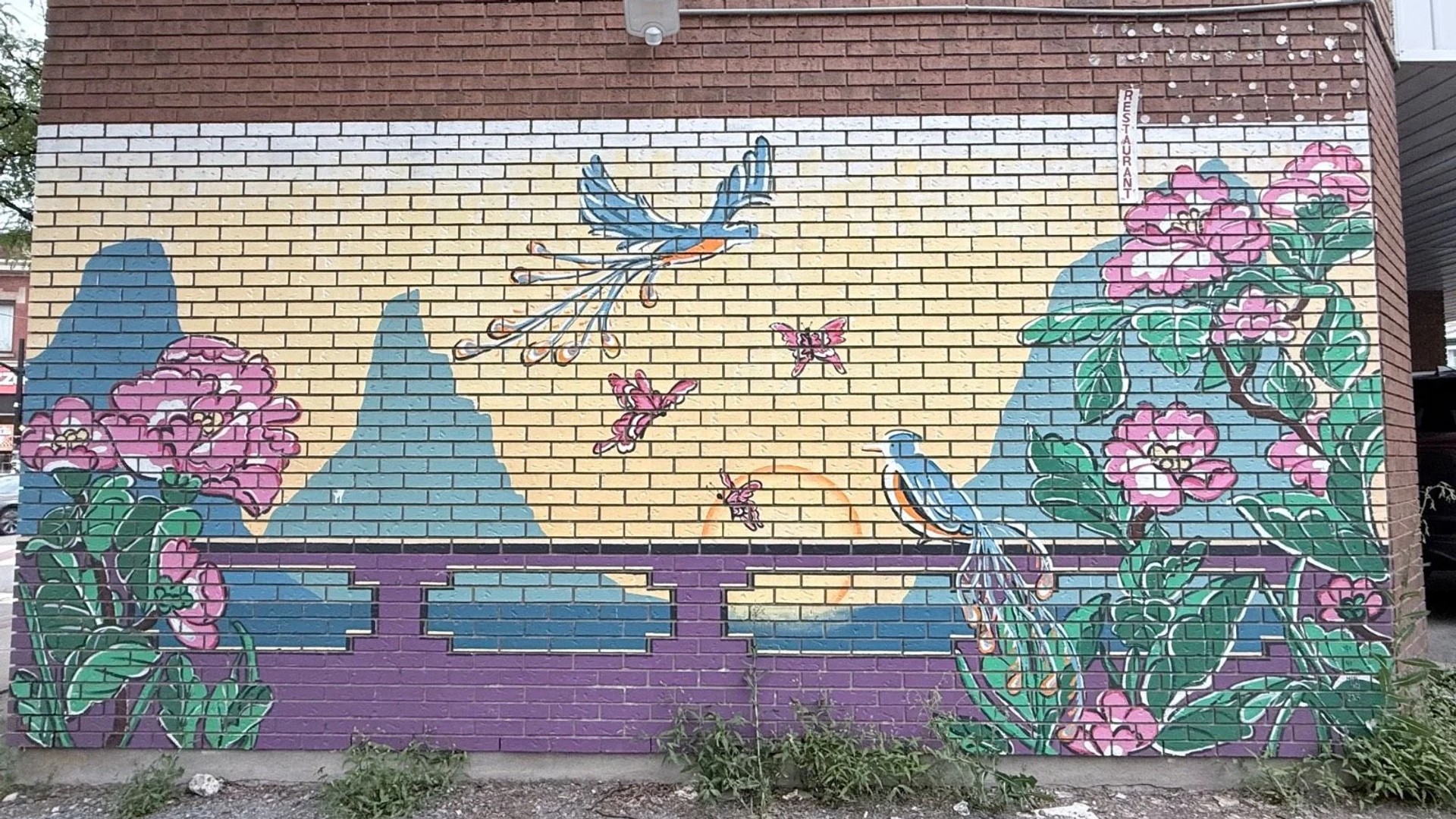 A mural on a brick wall depicts a blue bird, pink butterflies, and large flowers against blue mountains at 299 Bronson Avenue, Chinatown, Ottawa.