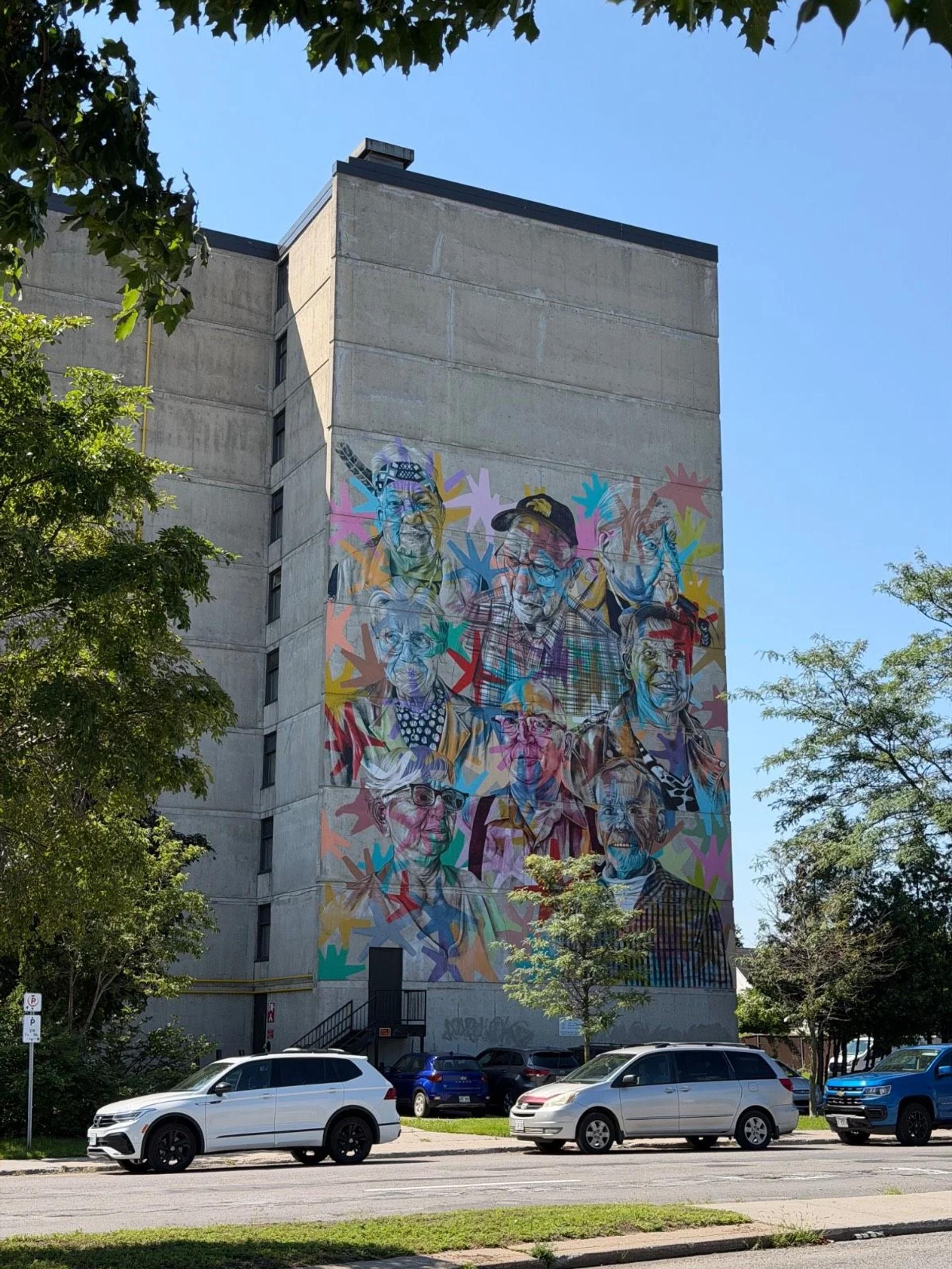 Vibrant pop-art mural of nine portraits on a concrete building, with cars below on Boulevard Sacré-Coeur, Hull, Gatineau, Ottawa.