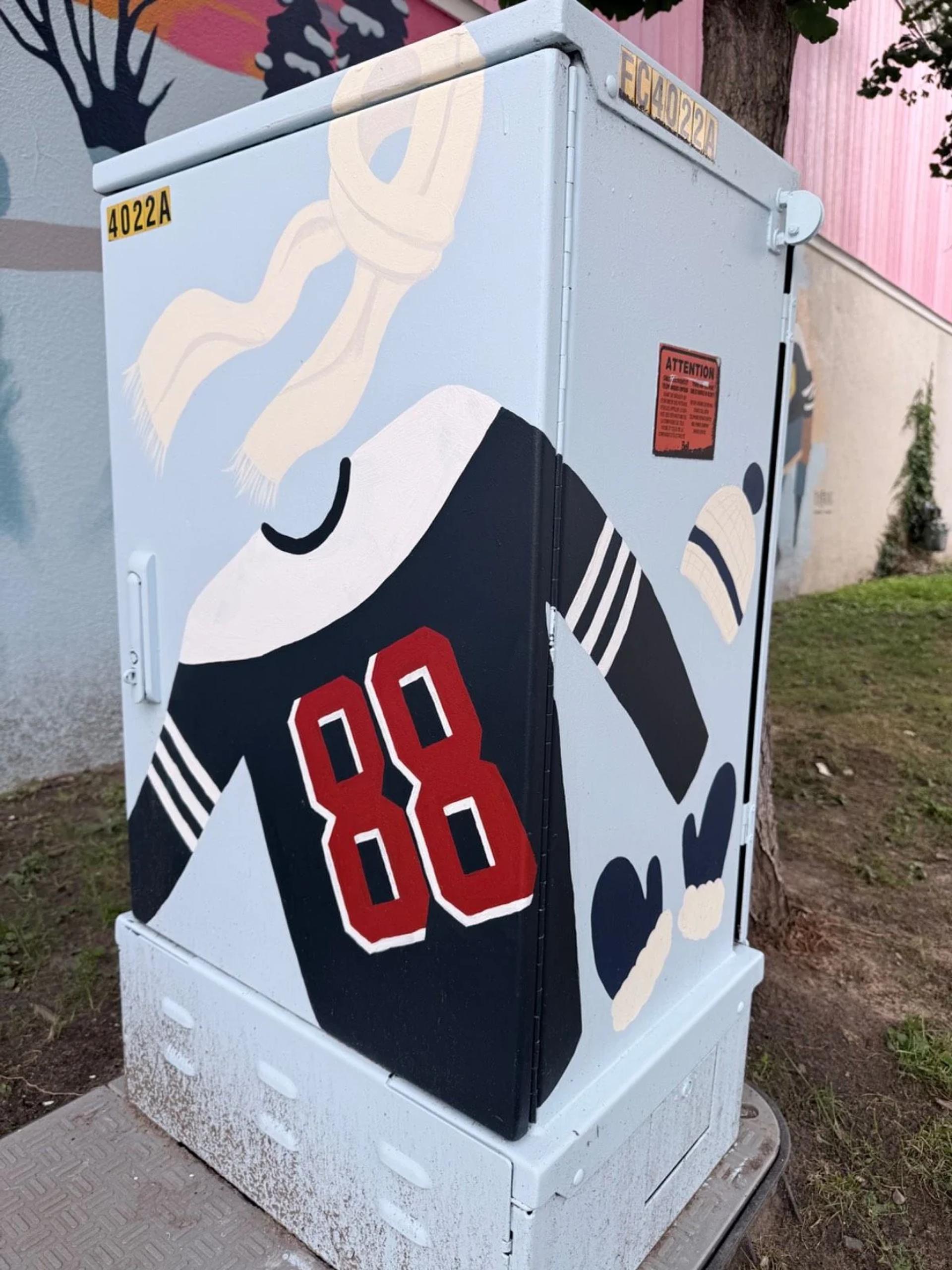 A light blue utility box painted with a cream scarf, dark blue jersey with 88, a knit hat, and mittens, on Bank Street, Ottawa.