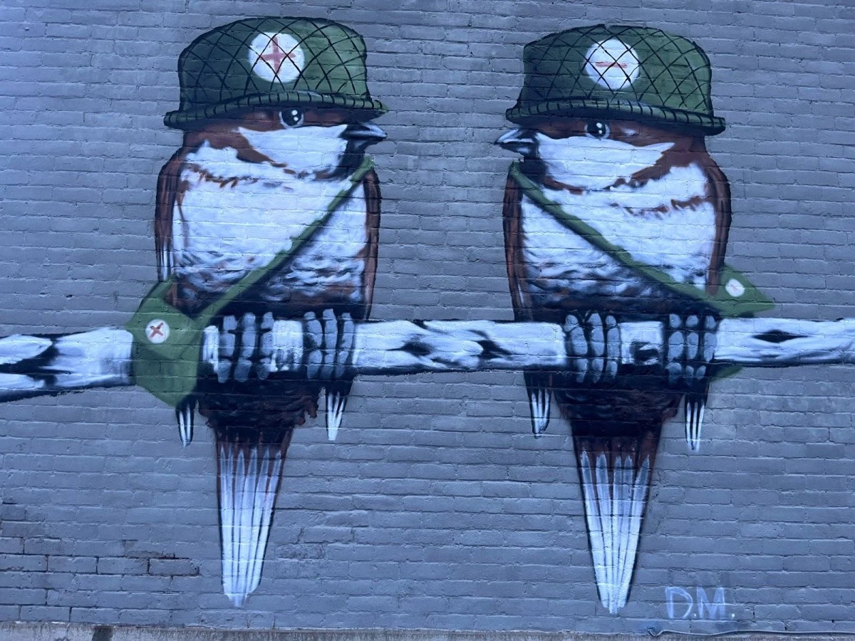 A brick wall mural by Daniel Martelock depicting two house sparrows wearing army helmets with plus and minus symbols, representing community and teamwork.