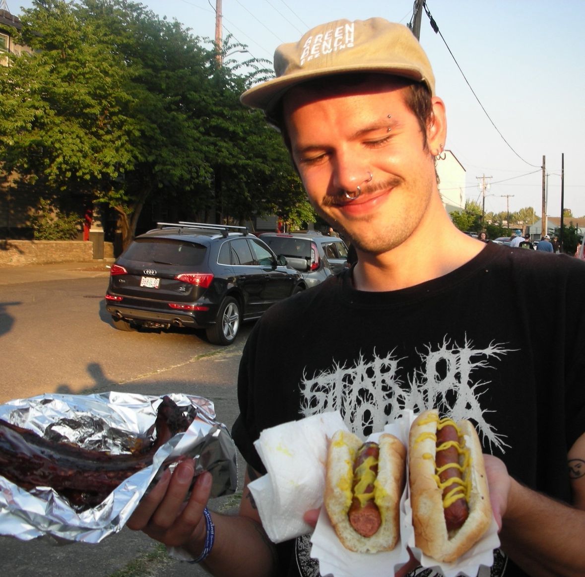 kurt holding 2 hot dogs with mustard and a bbq rib wearing a heavy metal t-shirt and a green bench brewing hat
