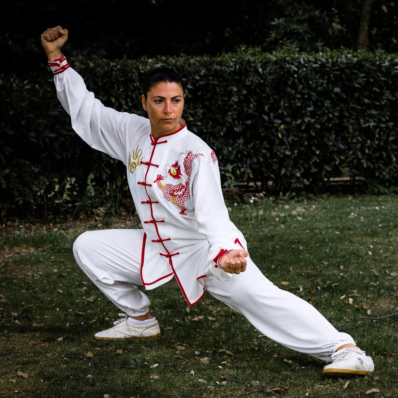 Una praticante in divisa bianca e rossa esegue una posizione bassa di Taiji Quan in un giardino.