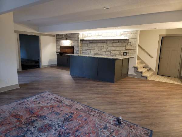 The remodeled basement in Overland Park, Kansas, with a granite topped wet bar, dark cabinets & a stone wall with a lit shelf