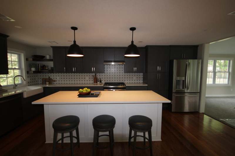 The kitchen seen end on, with the quartz island, three stools, black dome pendants & the refrigerator set into charcoal cabinetry