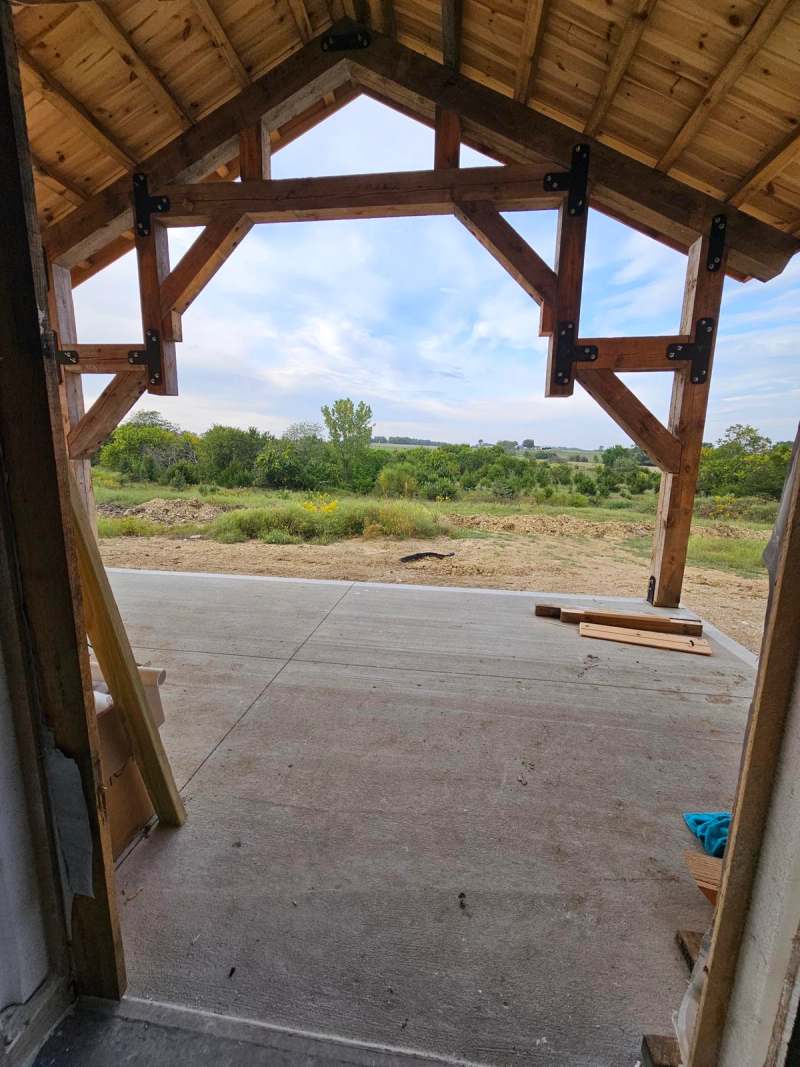 Standing under the covered porch, looking out through the timber truss to the open ground beyond
