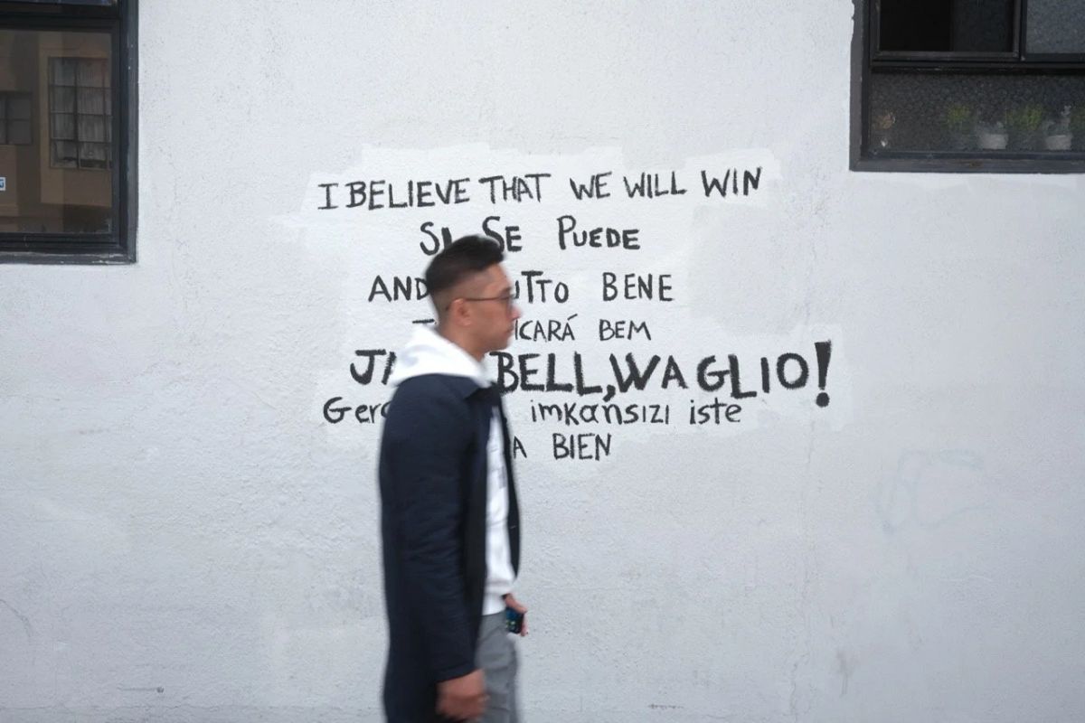 A man walks past a white wall painted with multilingual phrases like "I believe that we will win"
