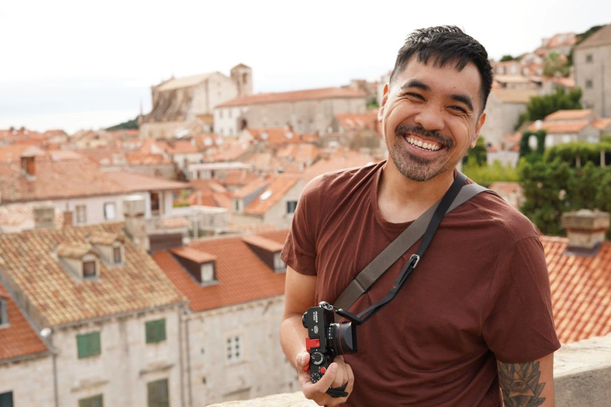 A smiling man with a camera on a strap stands above the terracotta rooftops of a Croatian old town