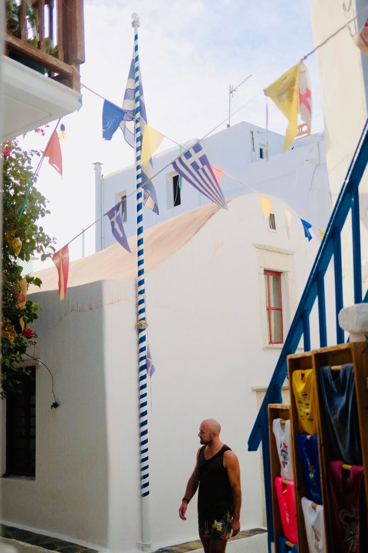 A man walks beneath strings of Greek flags and a blue-and-white striped pole in a whitewashed alley