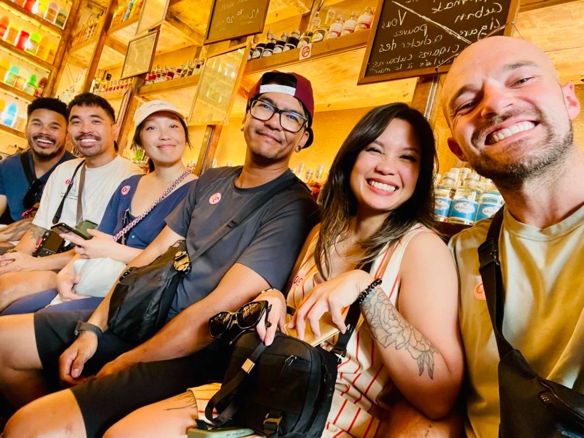 A group of six friends smiles for a selfie on a bench inside an ouzo bar lined with bottles