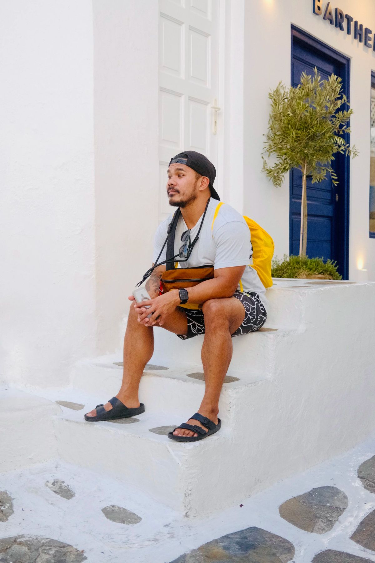 A man in a backwards cap sits on whitewashed steps beside a blue door and a potted olive tree