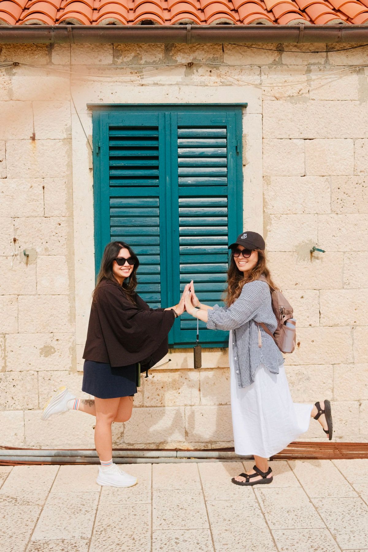Two women high-five in front of a green-shuttered window on a pale stone building with a tiled roof