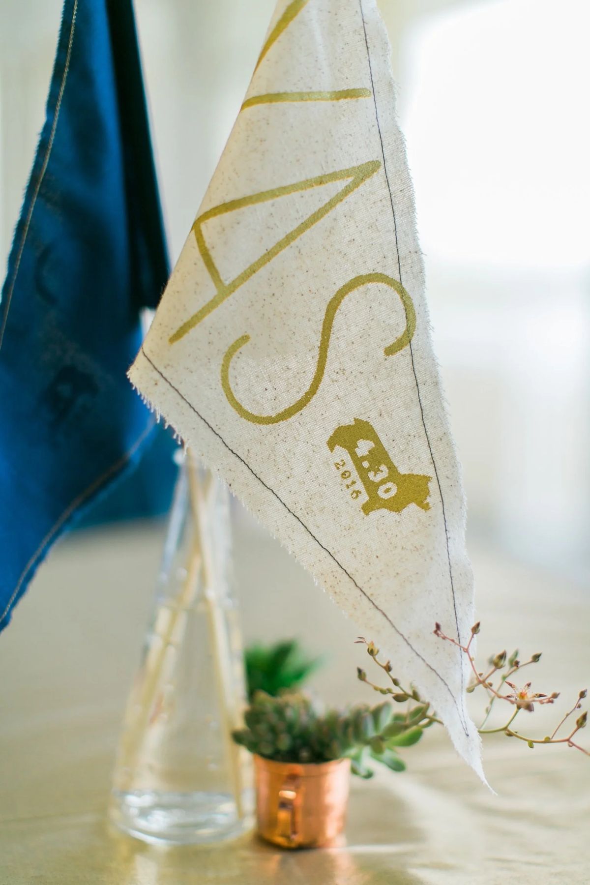 Cream fabric pennant printed in gold with "YAS" and a 4.30.2016 date stamp, beside a small succulent in a copper pot