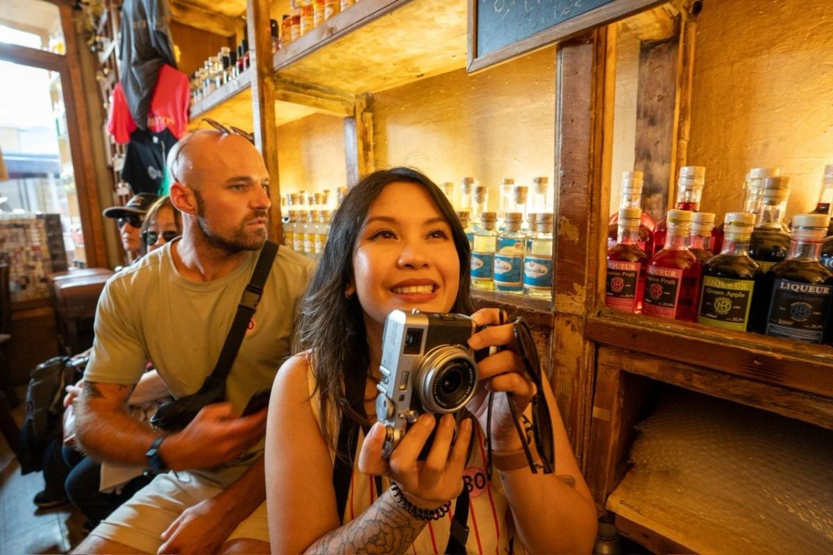 A woman smiles while holding a compact camera in an ouzo bar, with liqueur bottles on the shelves