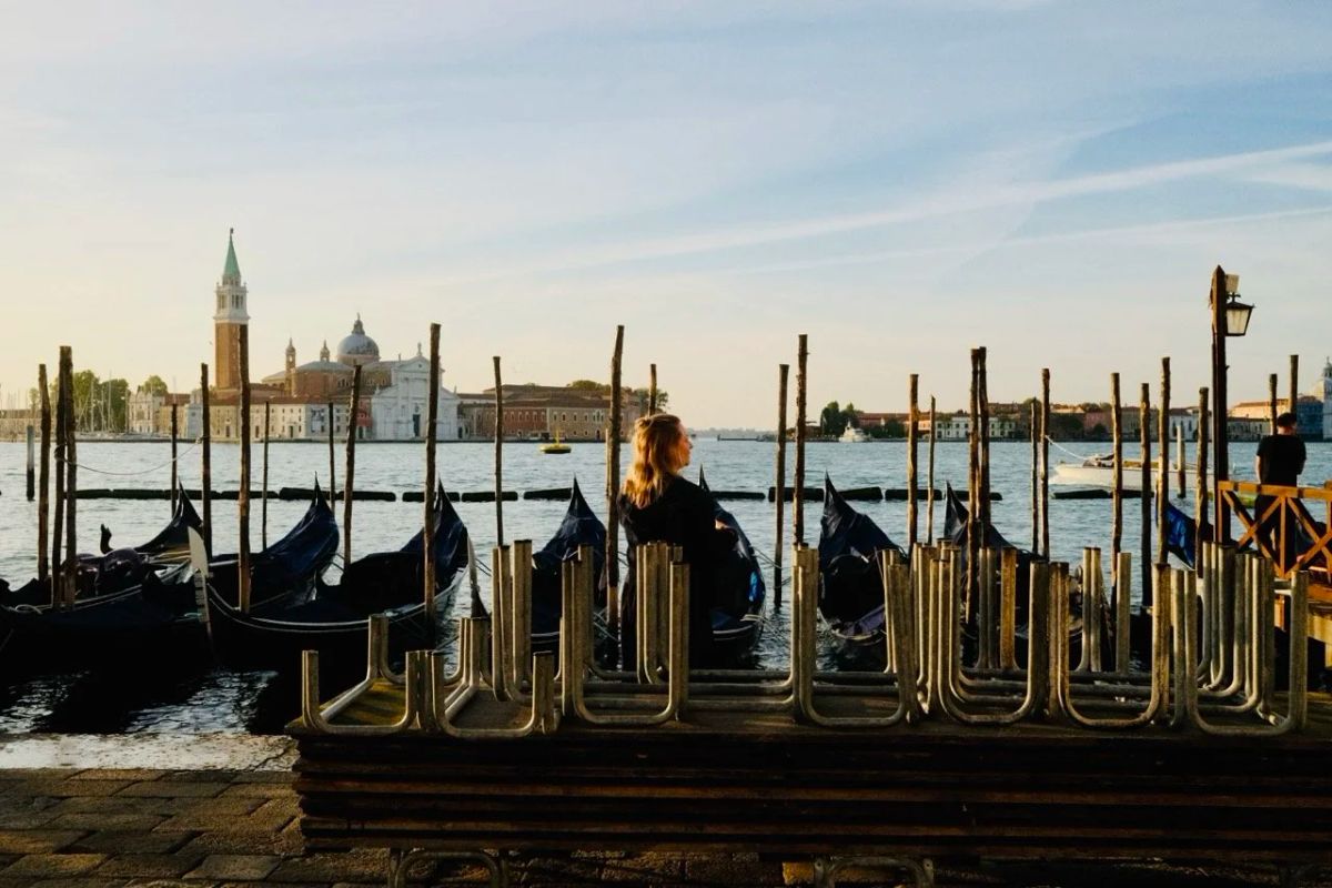 A woman sits on a waterfront bench among moored gondolas at sunset, San Giorgio Maggiore behind