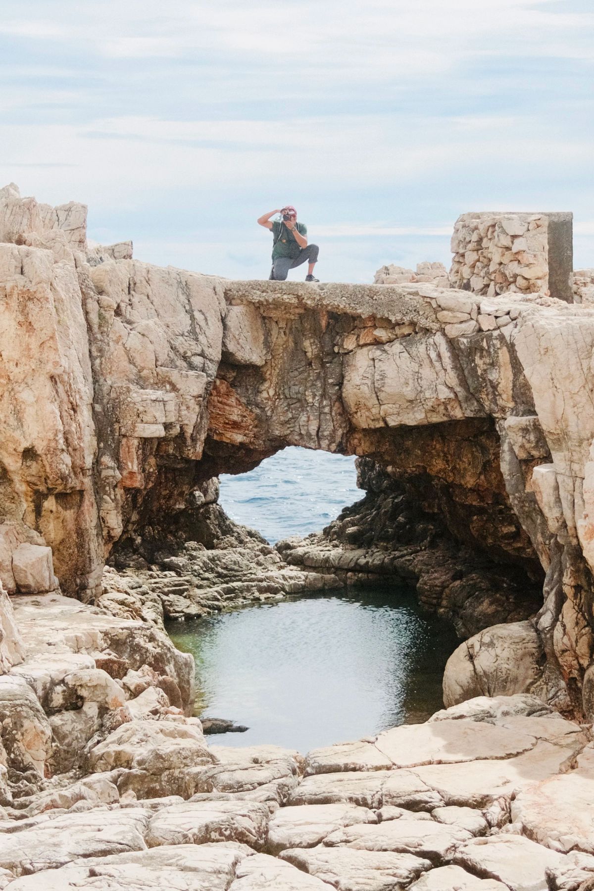 A person kneels to take a photo atop a natural rock arch above a tide pool on Lokrum island