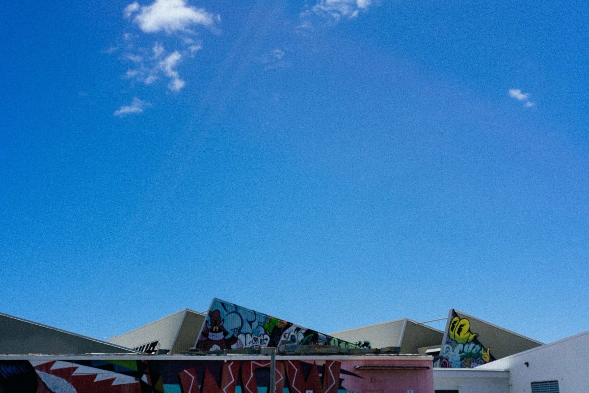 Wide blue sky over rooftops topped with colorful graffiti murals, a few wispy clouds above