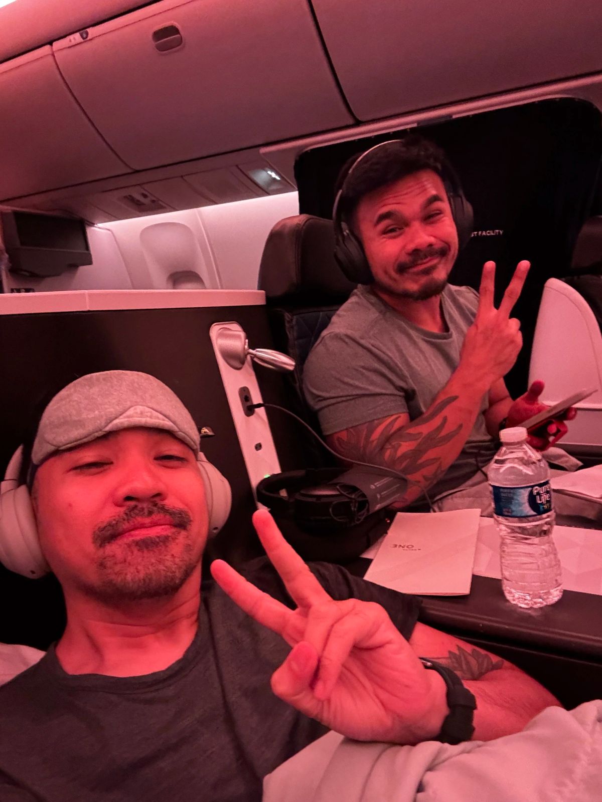 Two men in headphones flash peace signs from their seats in a business class airplane cabin