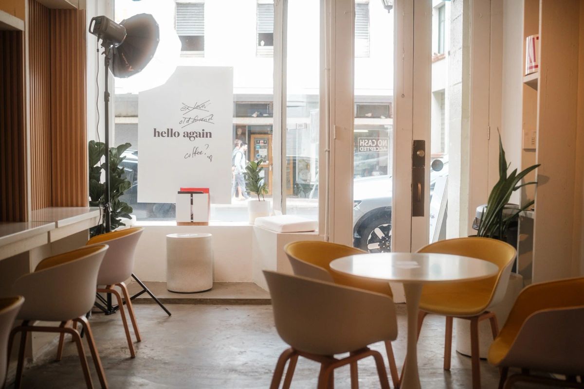 Bright cafe interior with mustard and white chairs, round tables and a "hello again" sign in the window