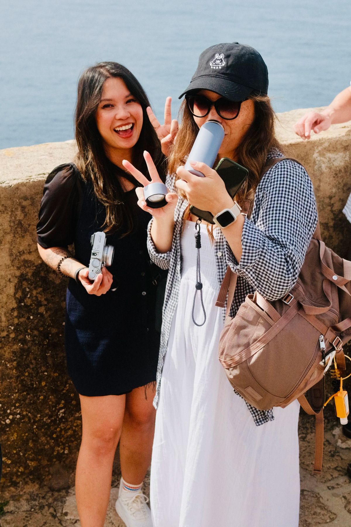 Two friends pose by a stone wall above the sea, one flashing a peace sign and one sipping from a bottle