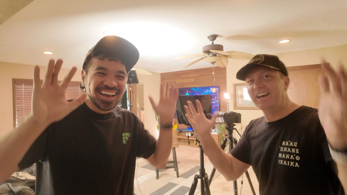 Two people on set waving and laughing at the camera, with a tripod, teleprompter and lights behind them, on location in Kailua, Hawaiʻi