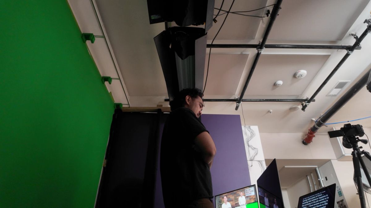 Low-angle view of a studio shoot: a person with folded arms watches the monitors beside a green screen, under a lighting grid, next to a teleprompter script, at a studio in San Diego