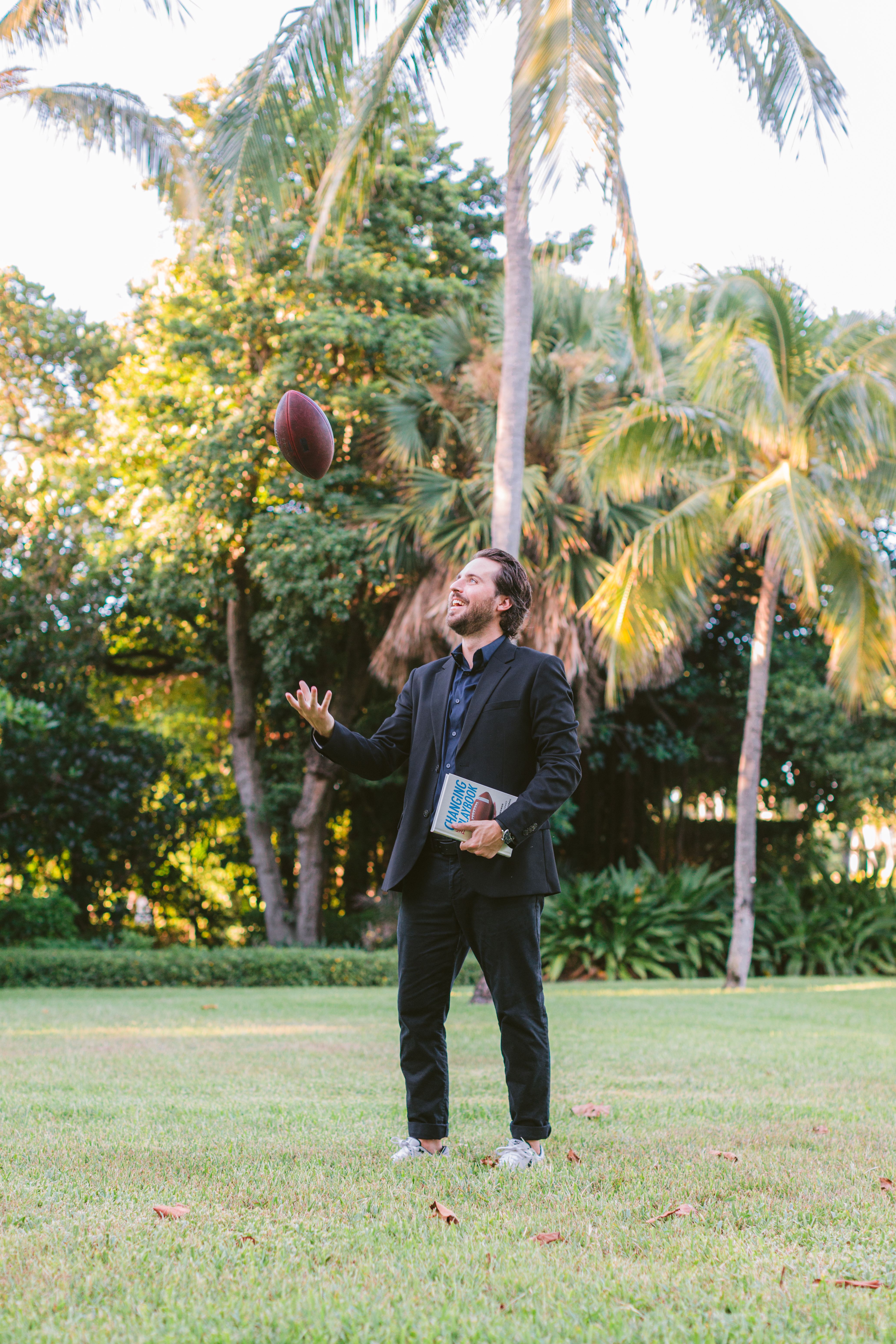Cash Lambert in a dark suit on a lawn under palm trees, holding a copy of Changing the Playbook and tossing a football in the air.