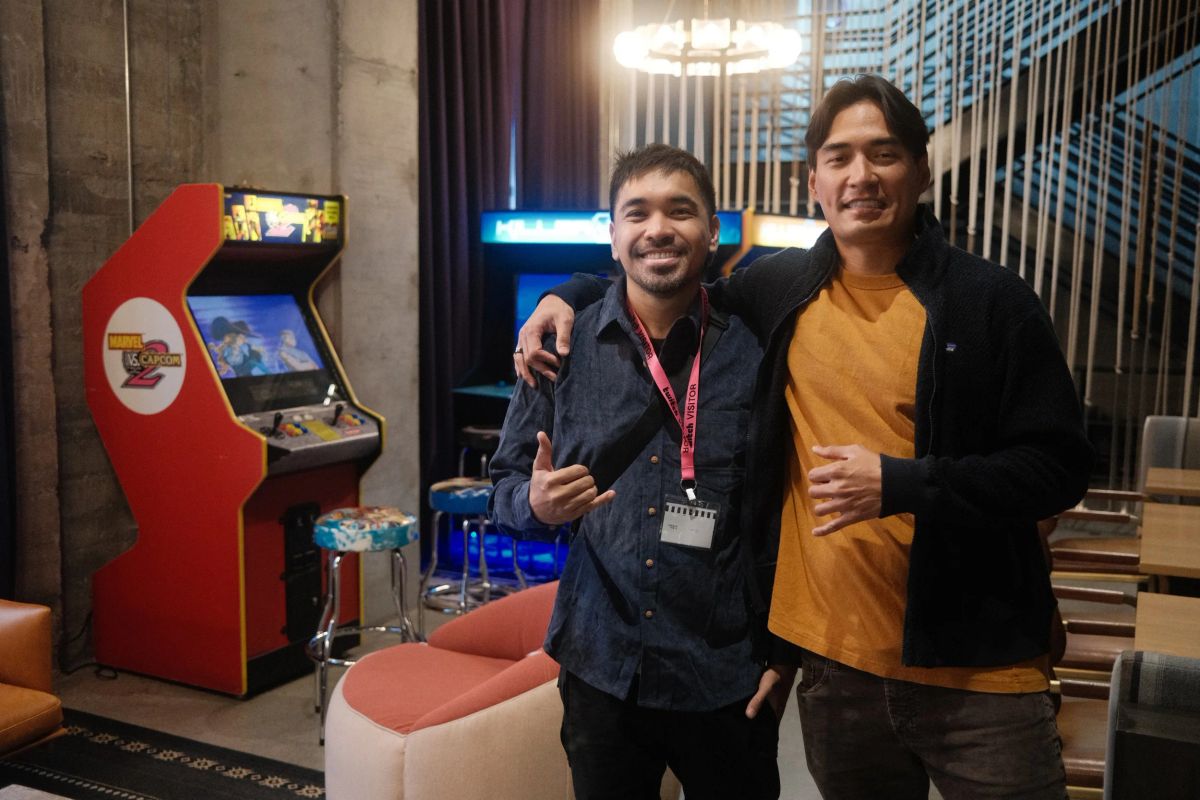 Two smiling men pose arm in arm in front of retro arcade cabinets in an office lounge