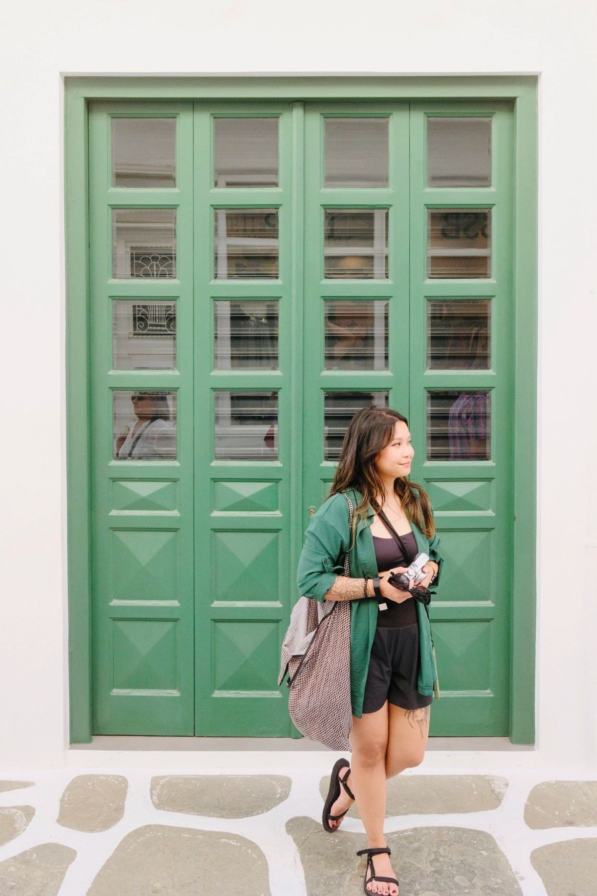A woman holding a camera stands before tall green paneled doors in a whitewashed Mykonos alleyway