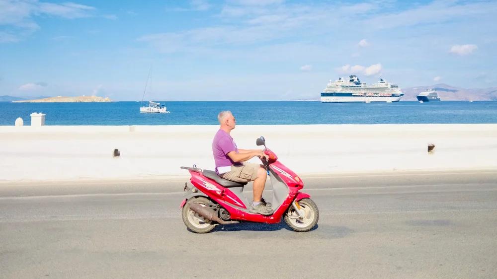 A man on a red scooter with cruise ships in the harbor behind him