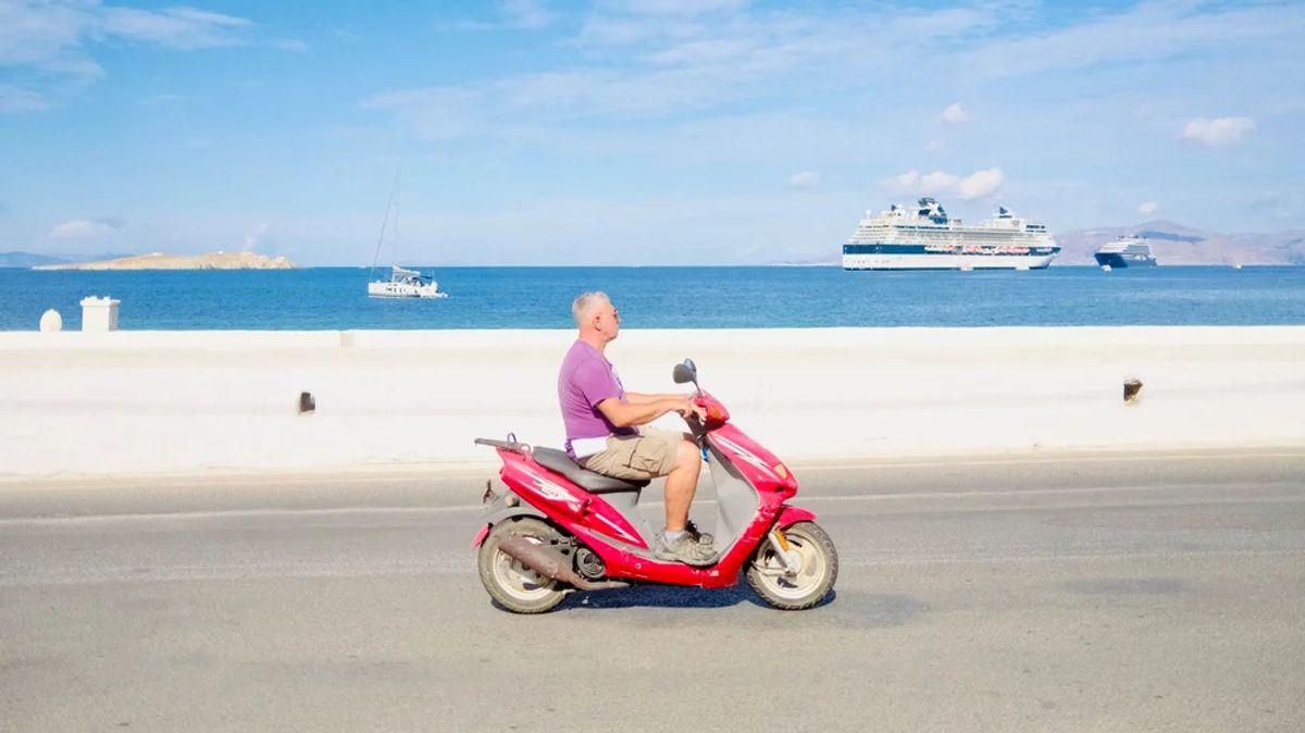 A man on a red scooter with cruise ships in the harbor behind him