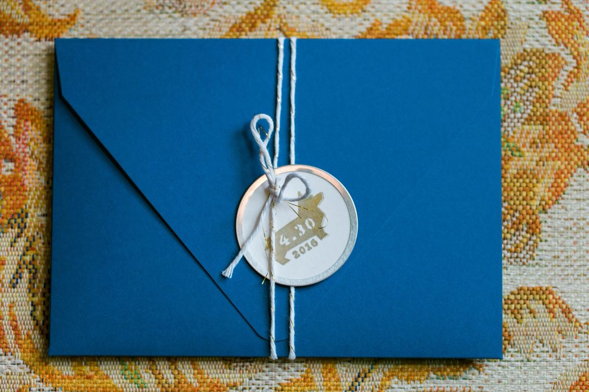 Blue wedding invitation envelope tied with white twine and a round 4.30.2016 seal, on a floral tapestry