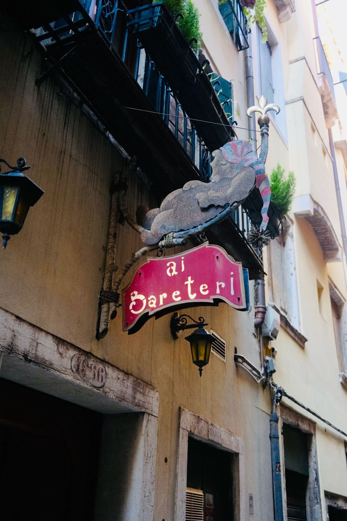 Red hanging sign reading "ai Bareteri" beneath a metal cat sculpture on a Venetian building facade