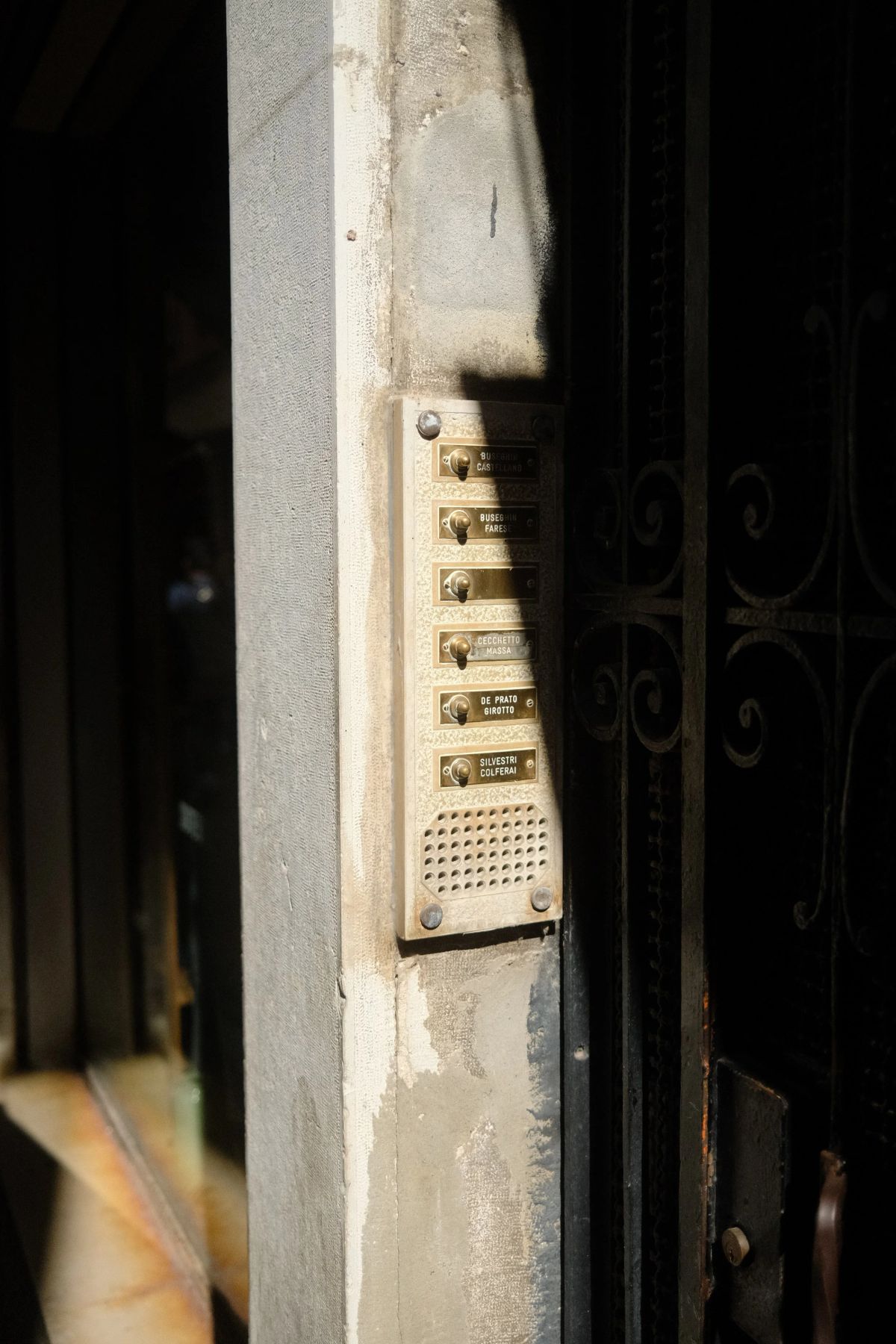 Sunlit brass doorbell panel with name buttons and a speaker grille beside an iron-gated doorway
