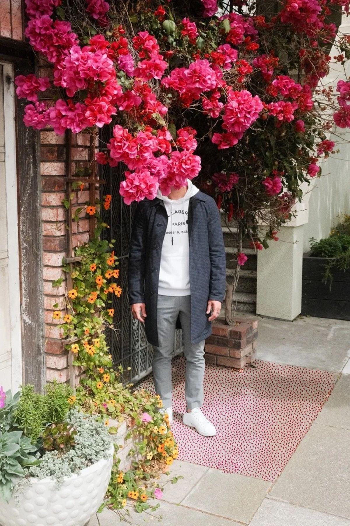 A man in a dark coat stands with his face hidden by a cascade of pink bougainvillea beside a doorway