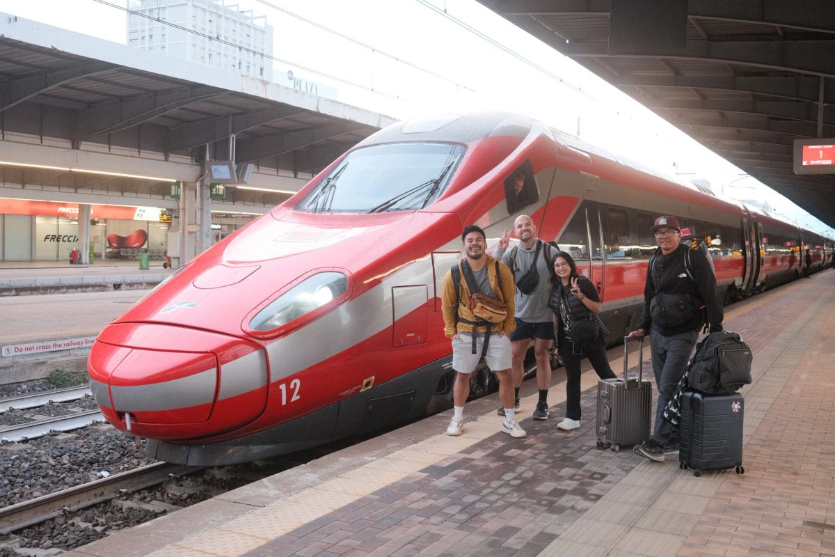 Four friends with luggage beside a red Frecciarossa high-speed train in Italy