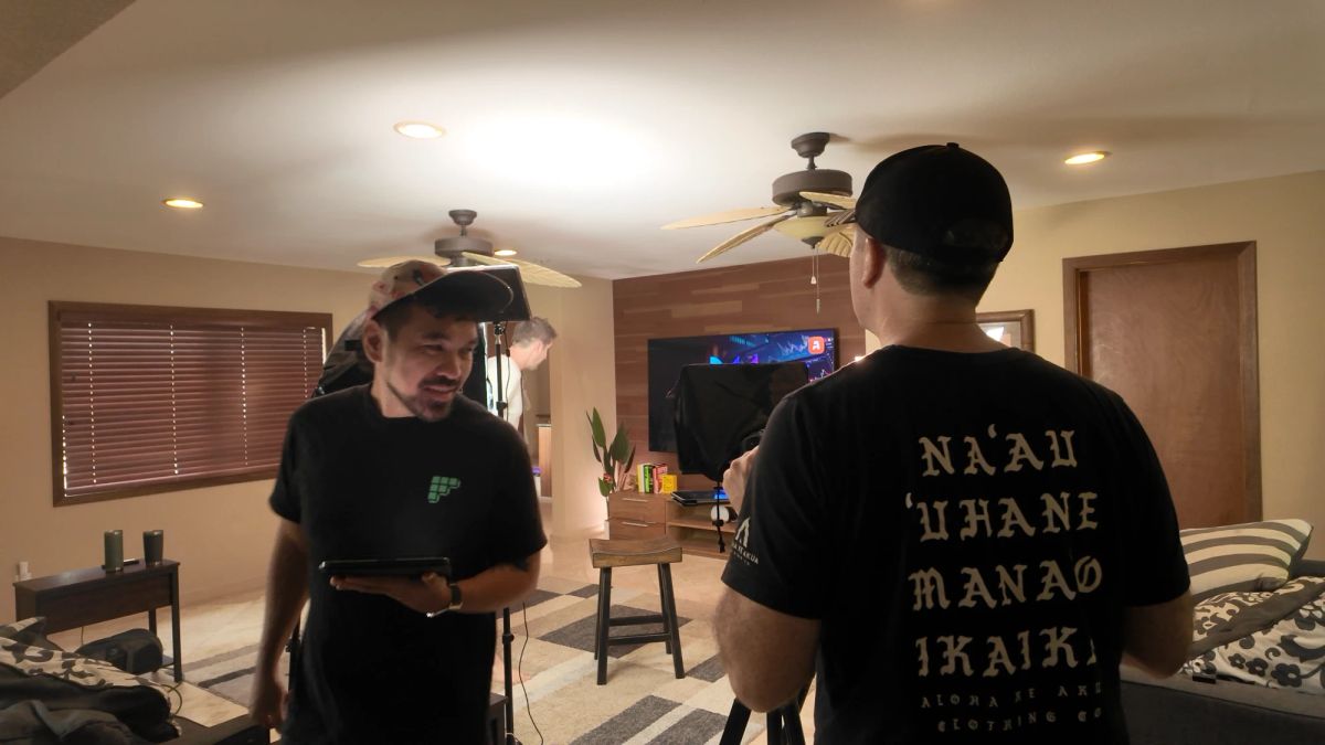 A living room turned into a set: one person holds a tablet while talking to another behind a camera and teleprompter, with an Awesomely screen on the TV, on location in Kailua, Hawaiʻi