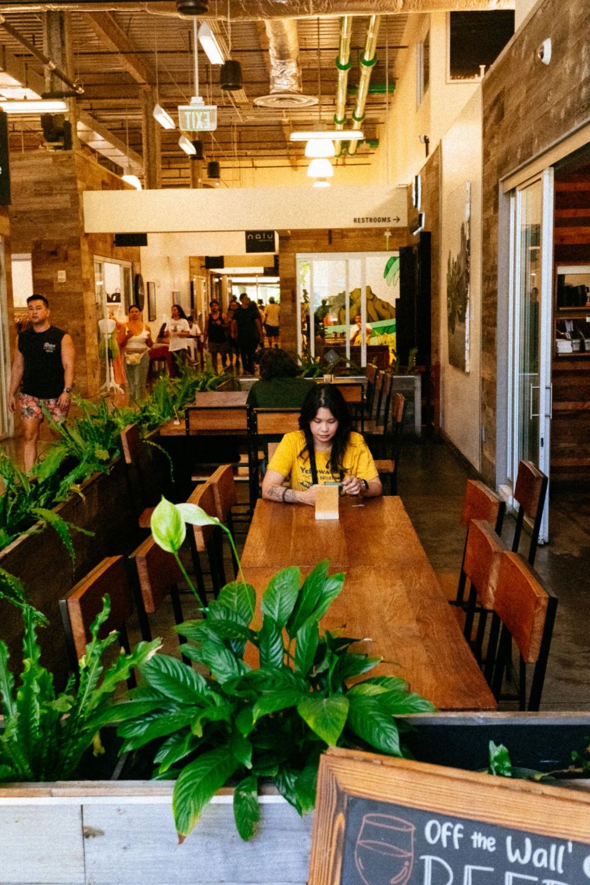 Woman in a yellow shirt seated at a long wooden table in a plant-filled food hall