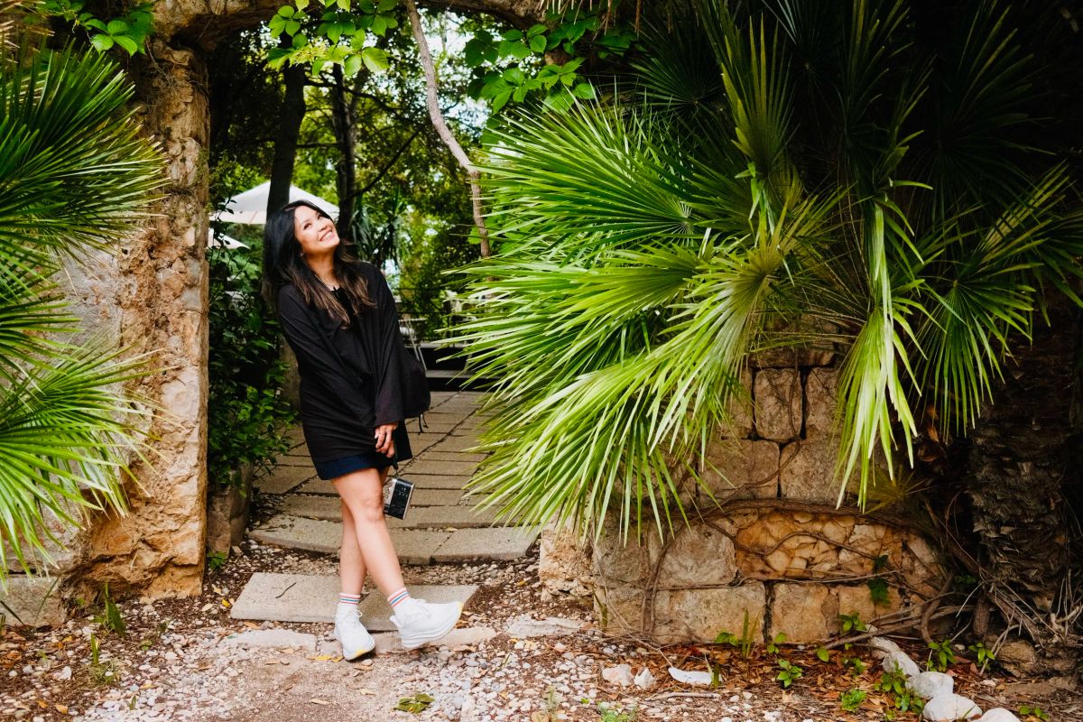 A smiling woman in black stands under a stone archway framed by large palm fronds