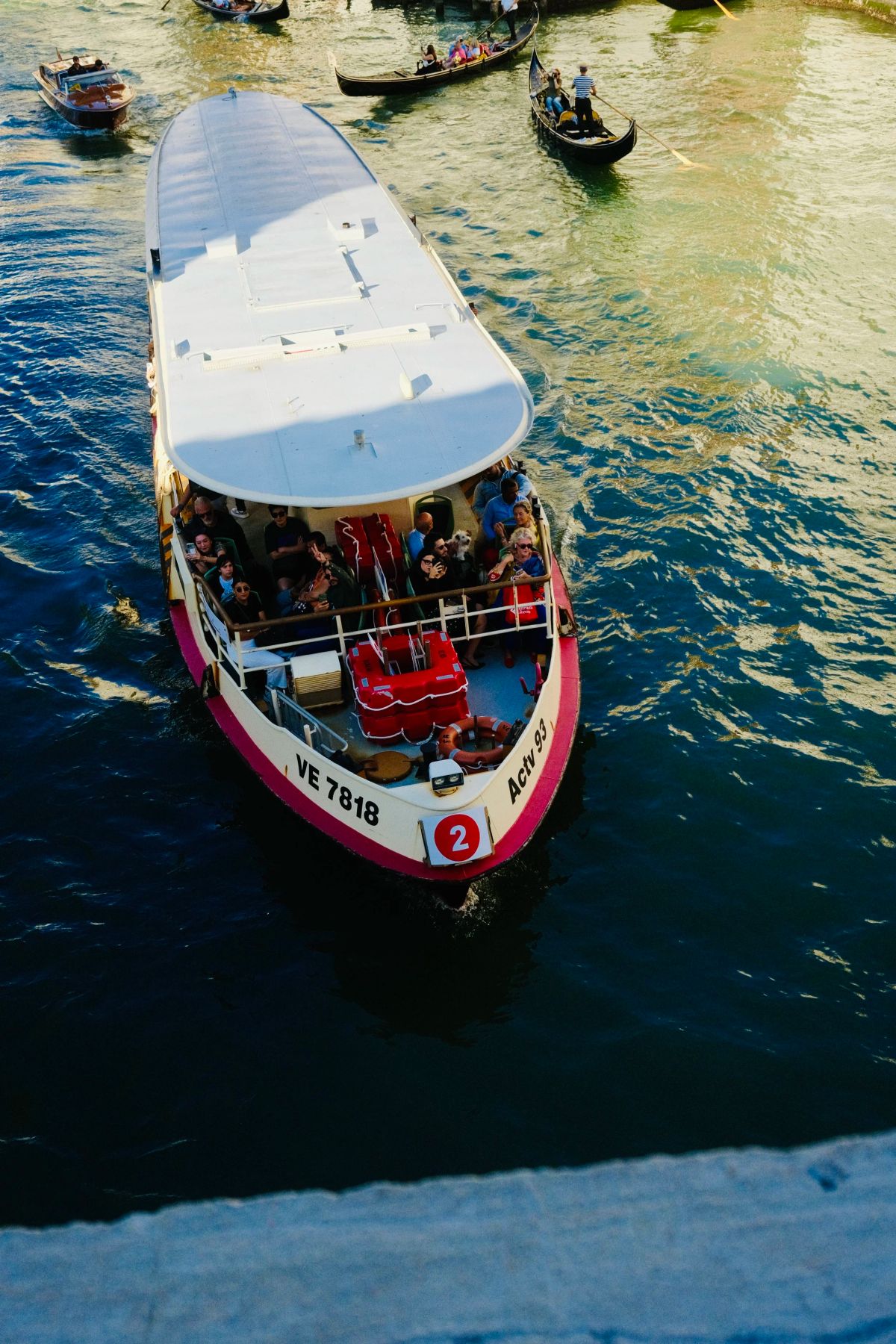 Overhead view of a red-and-white tour boat packed with passengers on a canal, gondolas nearby