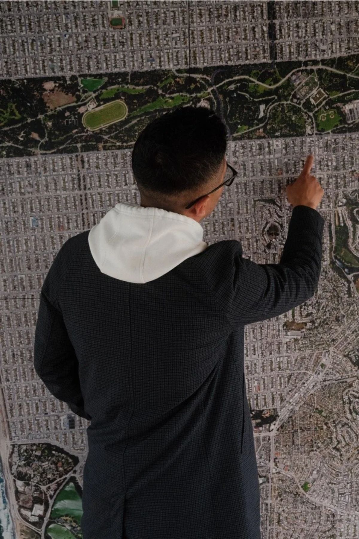 A man in a dark coat and white hoodie points at a large aerial city map covering a wall