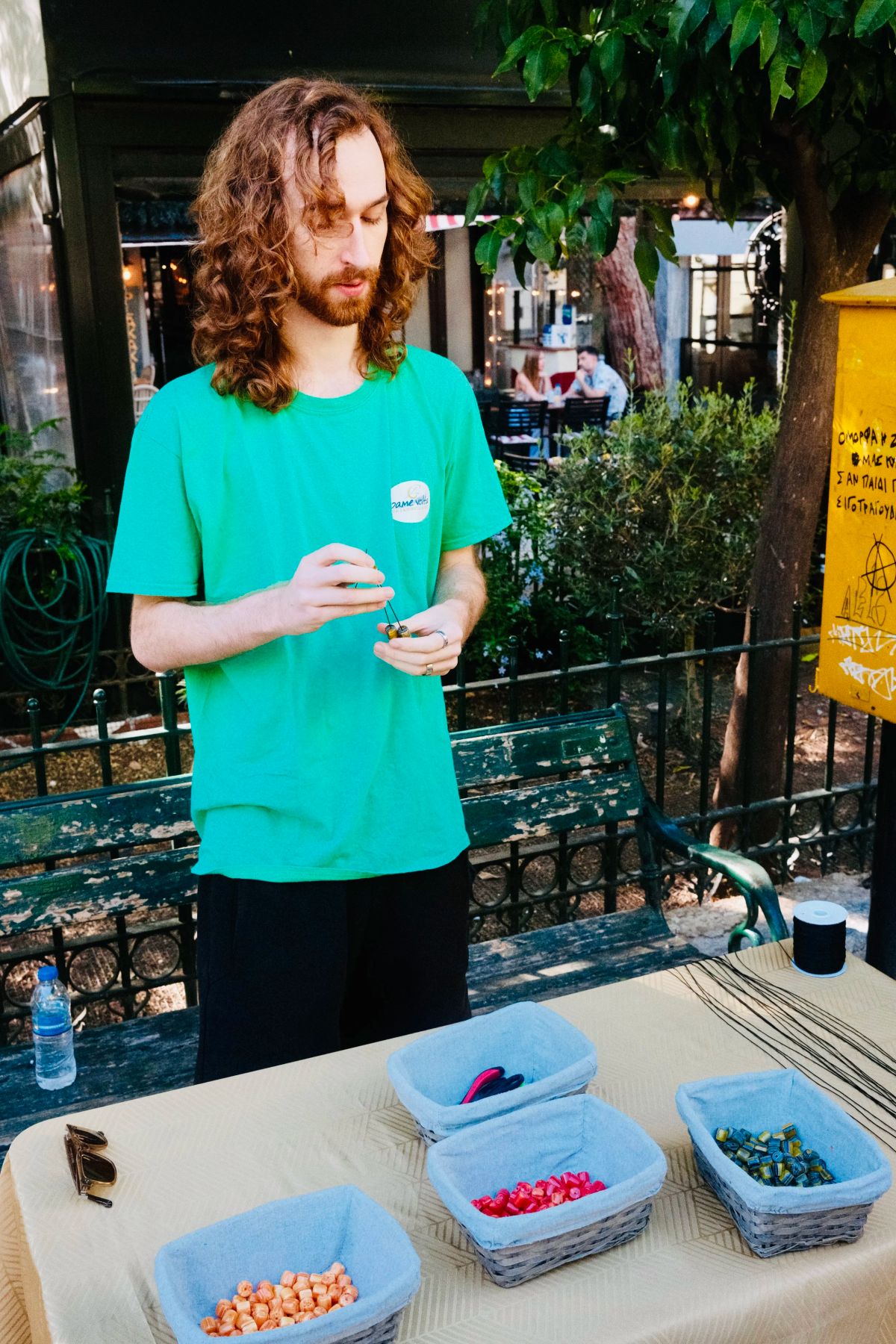 A man in a green shirt threads beads at a table of bead baskets during a komboloi workshop in Athens