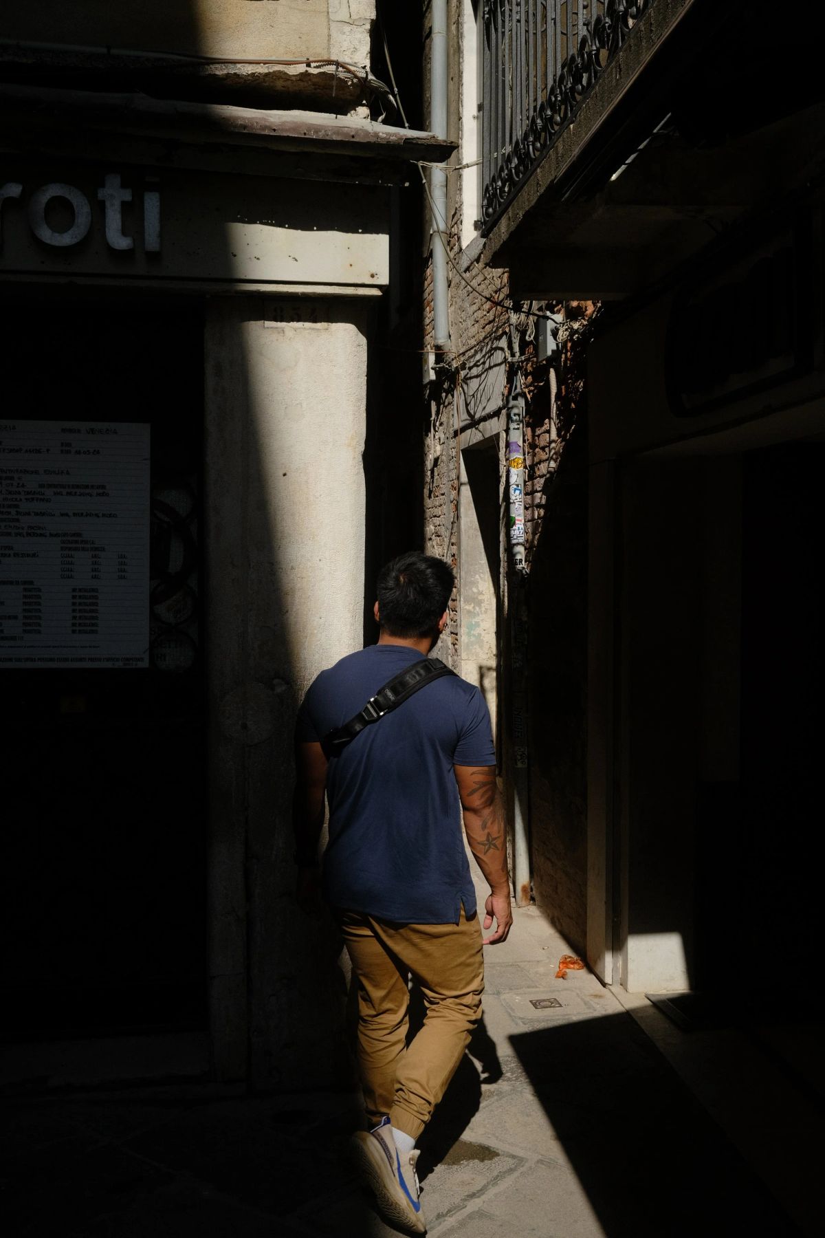 A man in a navy shirt walks into a narrow, shadowed Venice alley lit by a sharp slice of sunlight