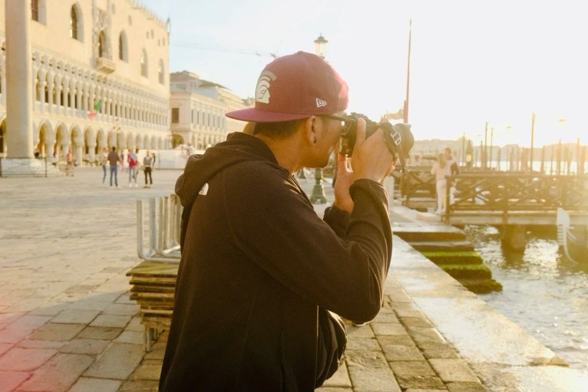 A man in a maroon cap photographs the Venice waterfront at golden hour beside the Doge's Palace