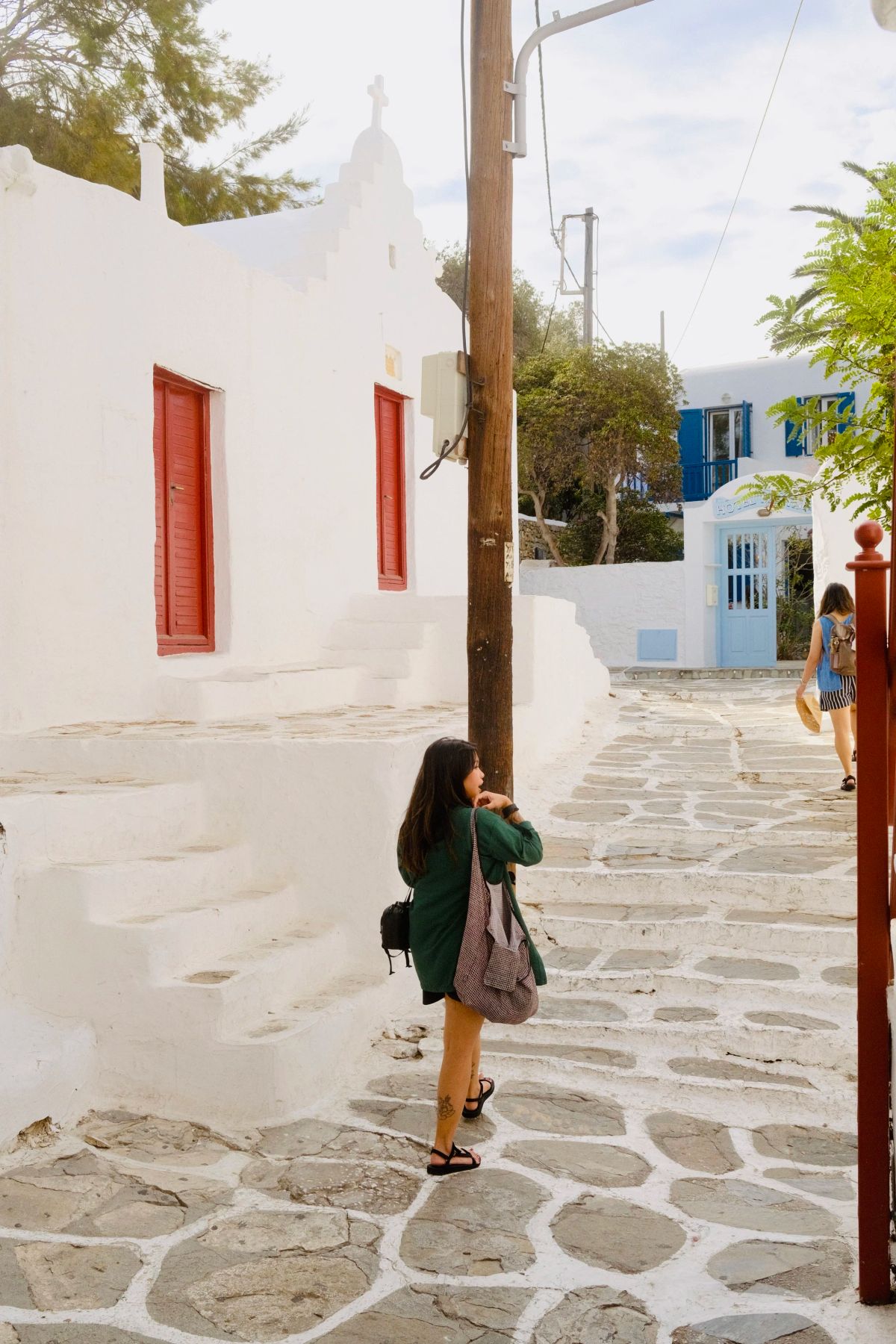 A woman in a green jacket walks up a whitewashed stone lane past a small chapel with red doors