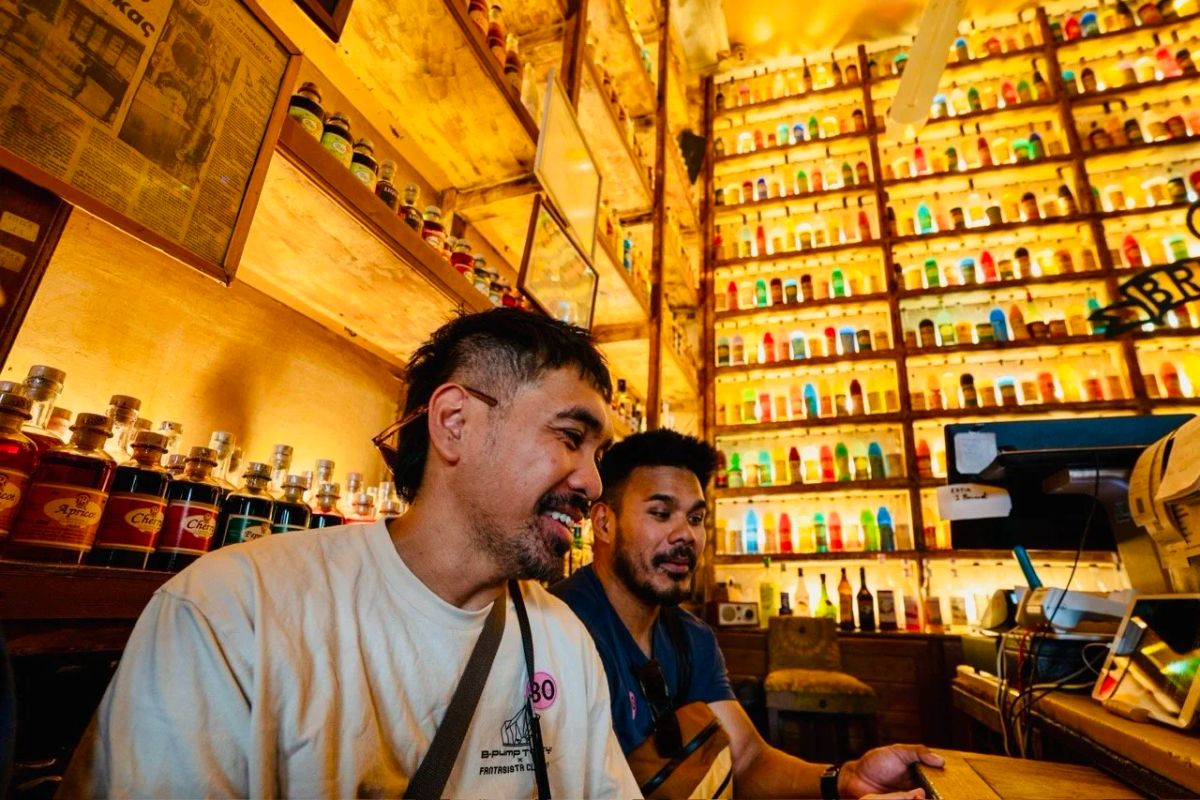 Two men laugh at the counter of a warmly lit ouzo bar with walls of colorful bottles behind them