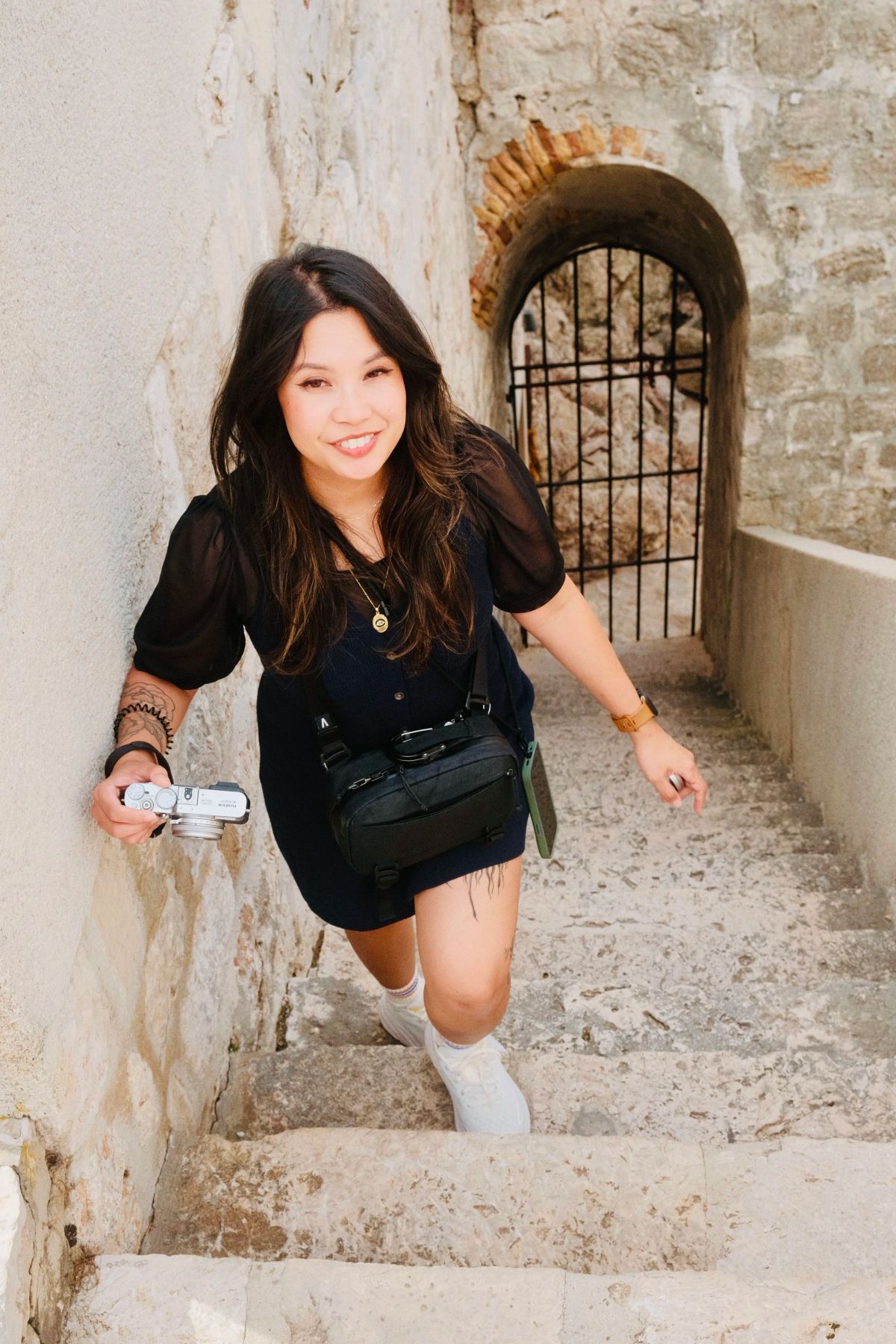 A smiling woman holding a small camera climbs worn stone steps toward an iron-gated archway