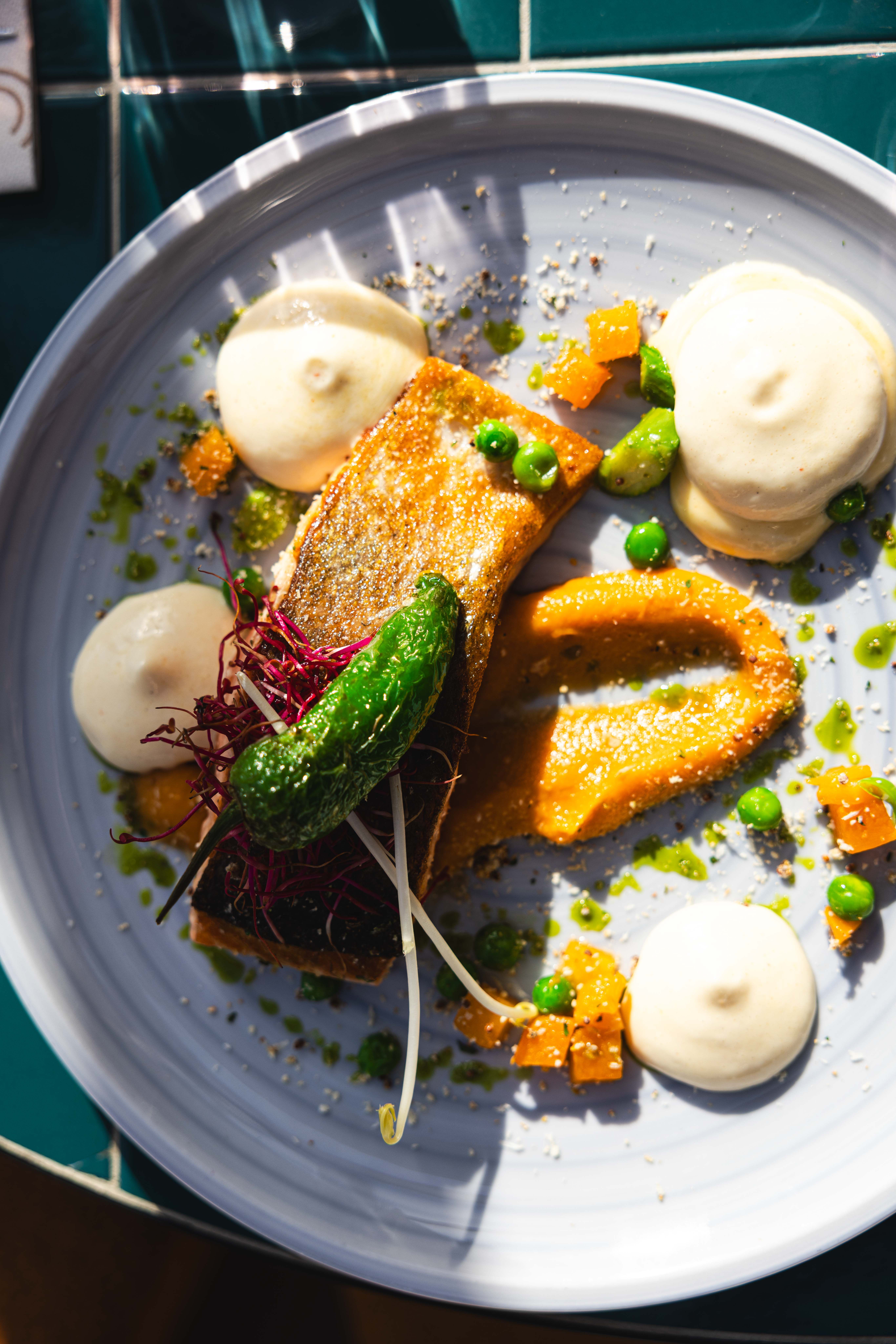 Gebakken visfilet met padrónpeper, pompoencrème en doperwten op een lichtblauw bord bij La Cocotte
