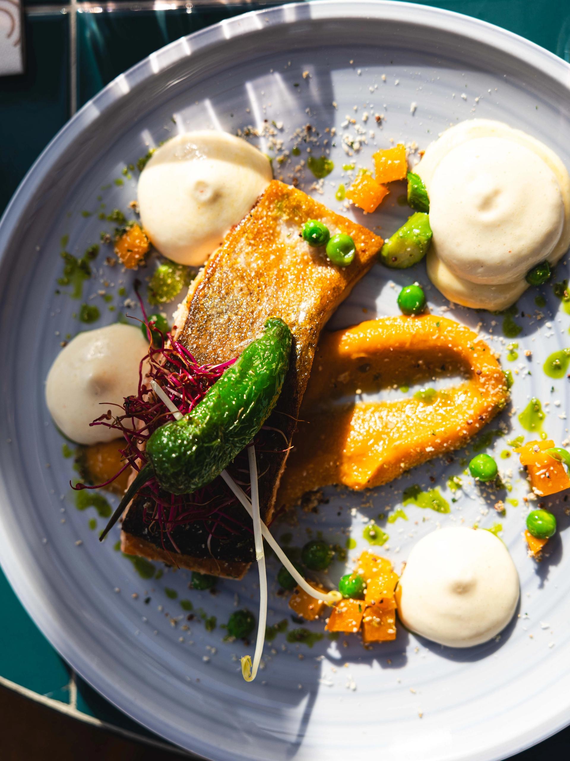Gebakken visfilet met padrónpeper, pompoencrème en doperwten op een lichtblauw bord bij La Cocotte